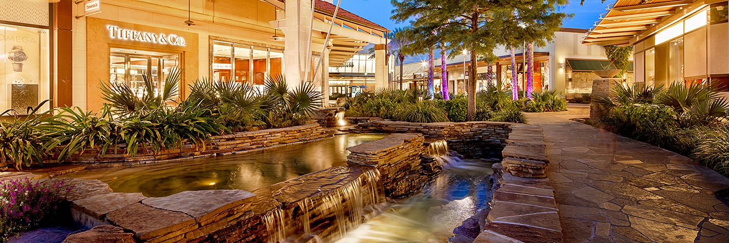 Tiered stone waterfalls in front of a Tiffany & Co. storefront at an upscale outdoor shopping center at twilight.