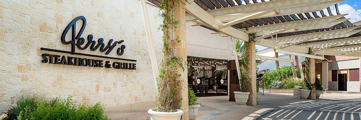Perry's Steakhouse & Grille sign on a light stone wall with a shaded pergola walkway and potted greenery.