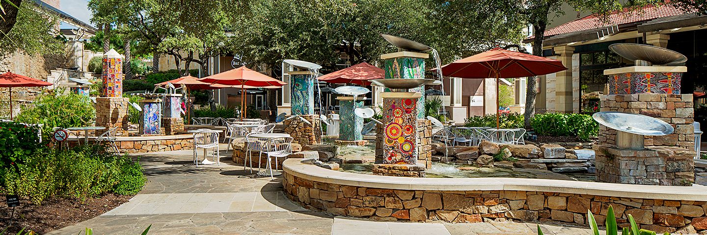 Stone patio with vibrant mosaic fountains, white dining tables, and red umbrellas surrounded by green trees.