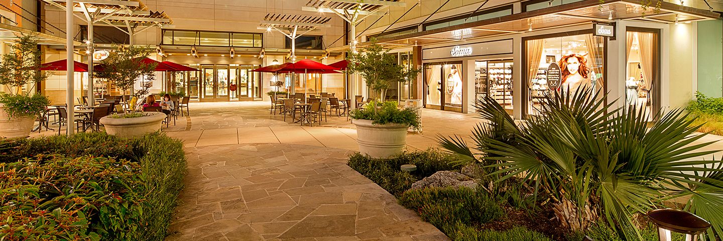 Illuminated Nordstrom and Soma stores at an outdoor shopping center with red umbrellas and manicured landscaping.