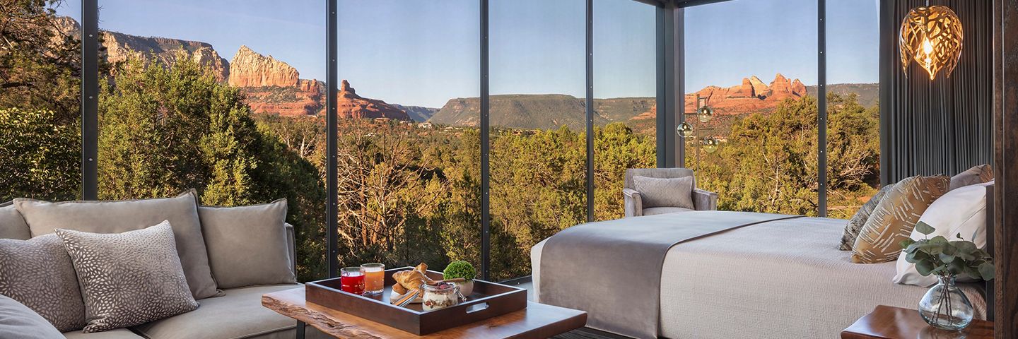 A modern bedroom with floor-to-ceiling glass walls offers a panoramic view of red rock formations and green trees in Sedona.