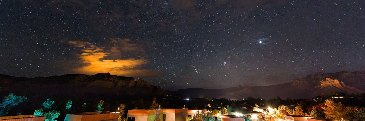A long-exposure night photograph shows a starry sky with a bright meteor streak over a darkened desert landscape and distant buildings.
