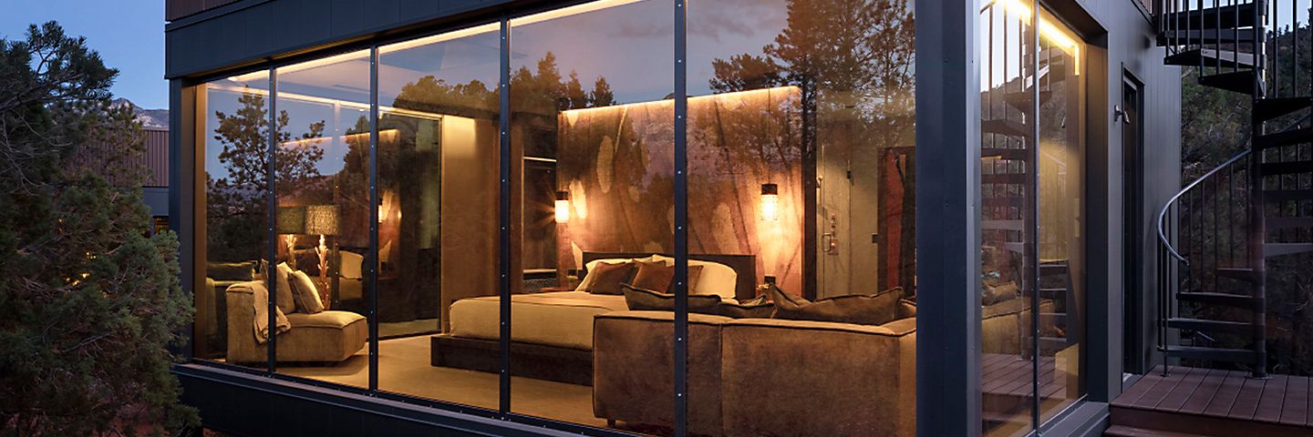 An exterior view through large glass windows reveals a luxury suite illuminated by warm interior lighting at dusk.