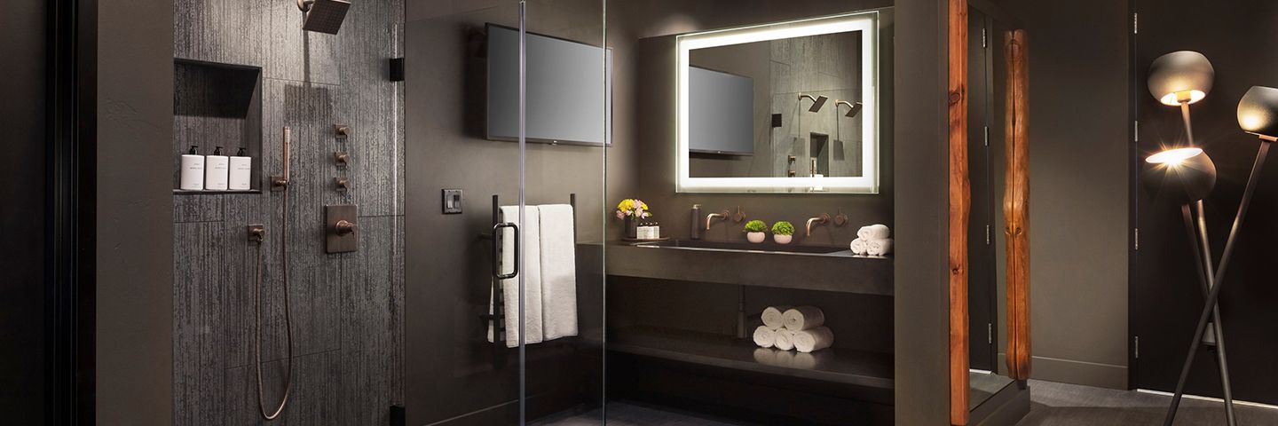 A contemporary bathroom features dark tiled walls, a large walk-in glass shower, and a minimalist vanity with a backlit mirror.