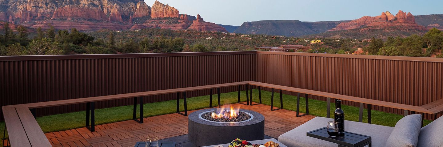 An outdoor rooftop patio includes a modern circular fire pit, wooden decking, and lounge seating against a mountain backdrop.