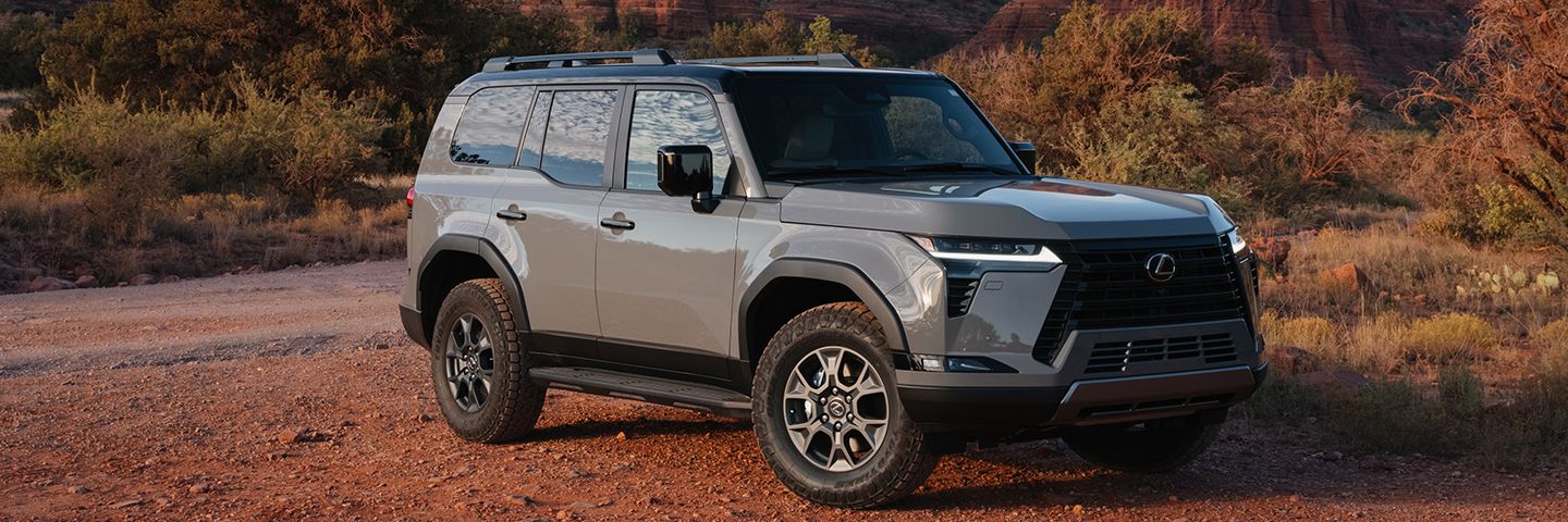 A grey Lexus SUV is parked on a dirt path in front of towering red sandstone cliffs under a clear sky.