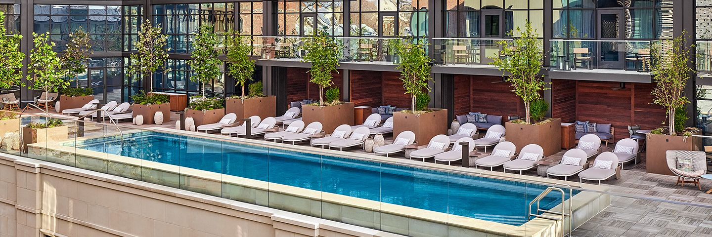 Luxury hotel pool deck featuring white loungers, greenery in large planters, and a multi-story glass-walled building.
