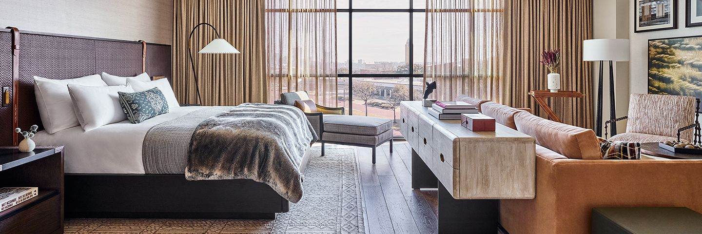 Modern hotel bedroom with a plush bed, chaise lounge, and floor-to-ceiling windows showing a city view.