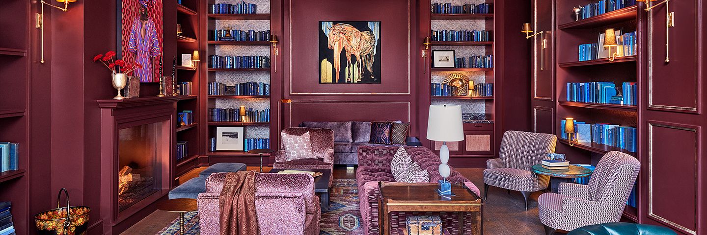 Luxurious library with burgundy walls, built-in shelves of blue books, purple velvet seating, and a fireplace.