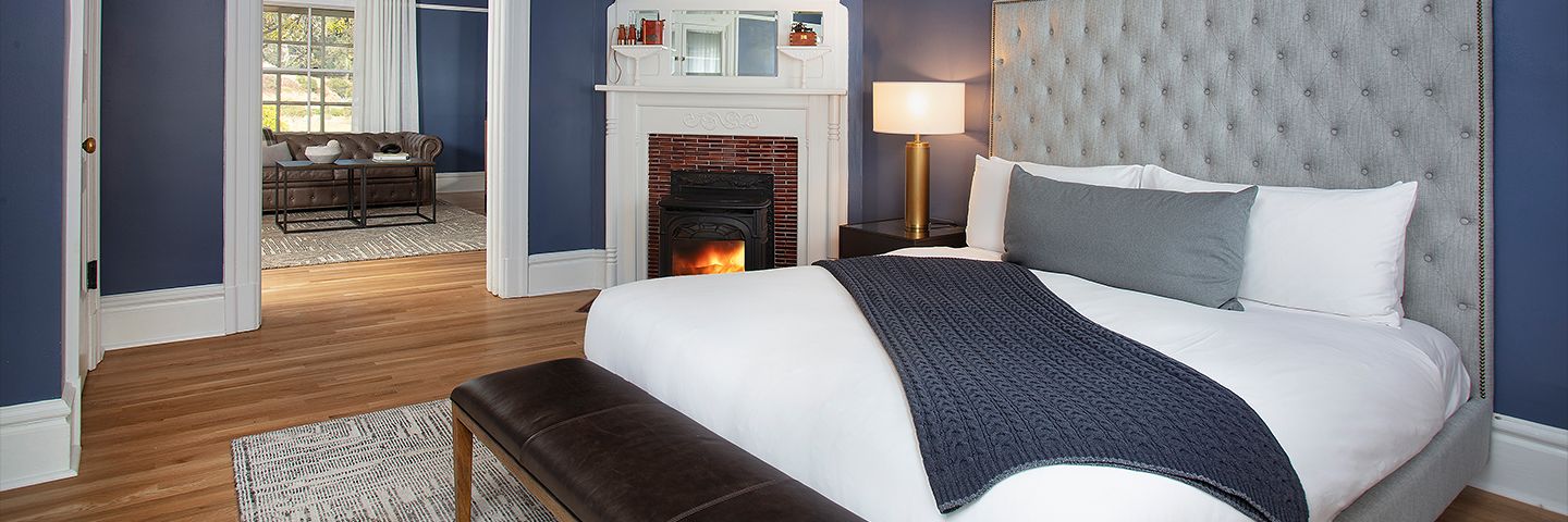 Modern navy bedroom with a tufted headboard, white fireplace with a lit stove, and view into an adjacent sitting area.