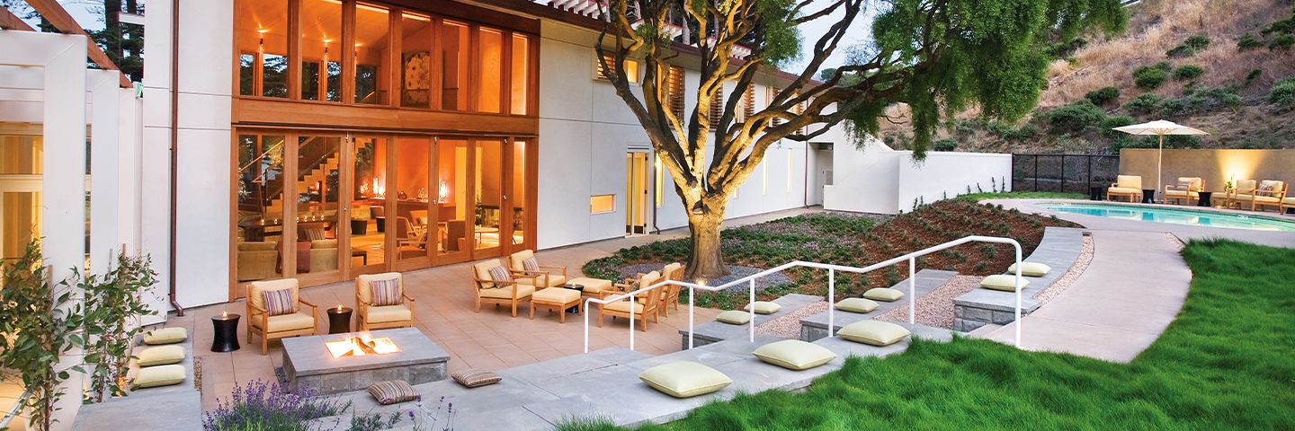 Modern home at dusk with a large glass facade, patio with fire pit, illuminated tree, and swimming pool.