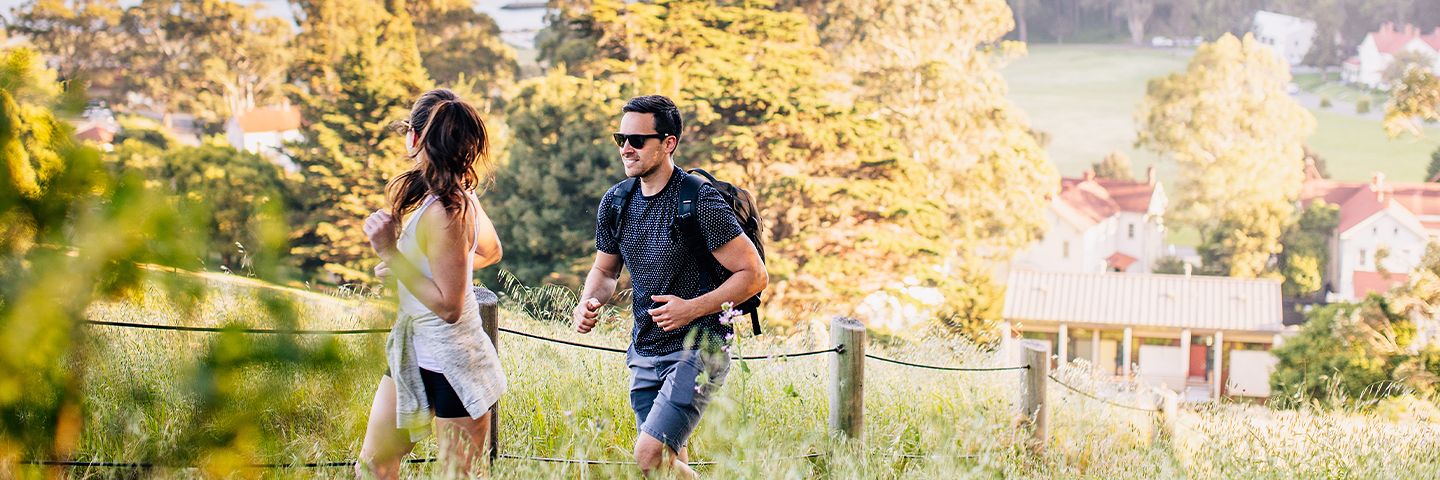 Man and woman jogging on a grassy hillside path overlooking a coastal bay and distant bridge.