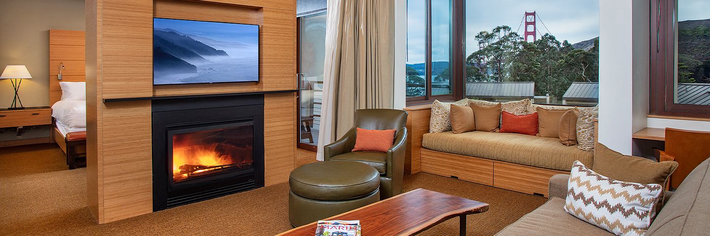 Modern hotel suite living room with a fireplace, TV, and a large window framing the Golden Gate Bridge.