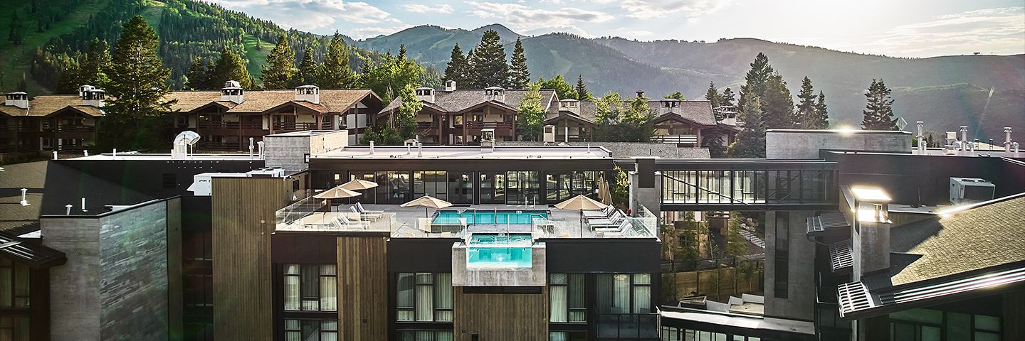 Modern resort rooftop with a pool and glass hot tub overlooking lush green mountains and pine forests.