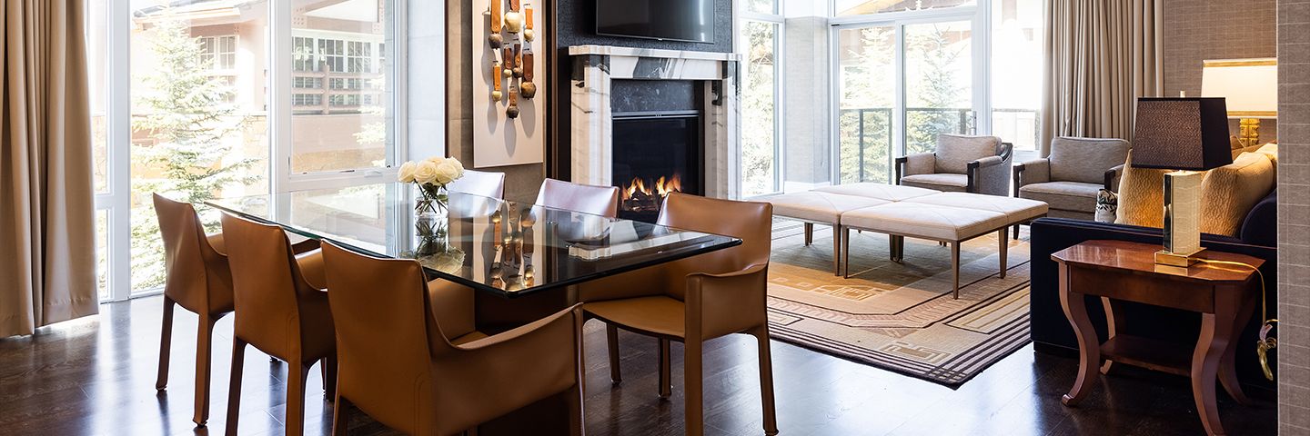 Modern open-concept living and dining room with a fireplace, glass table, tan leather chairs, and large windows.
