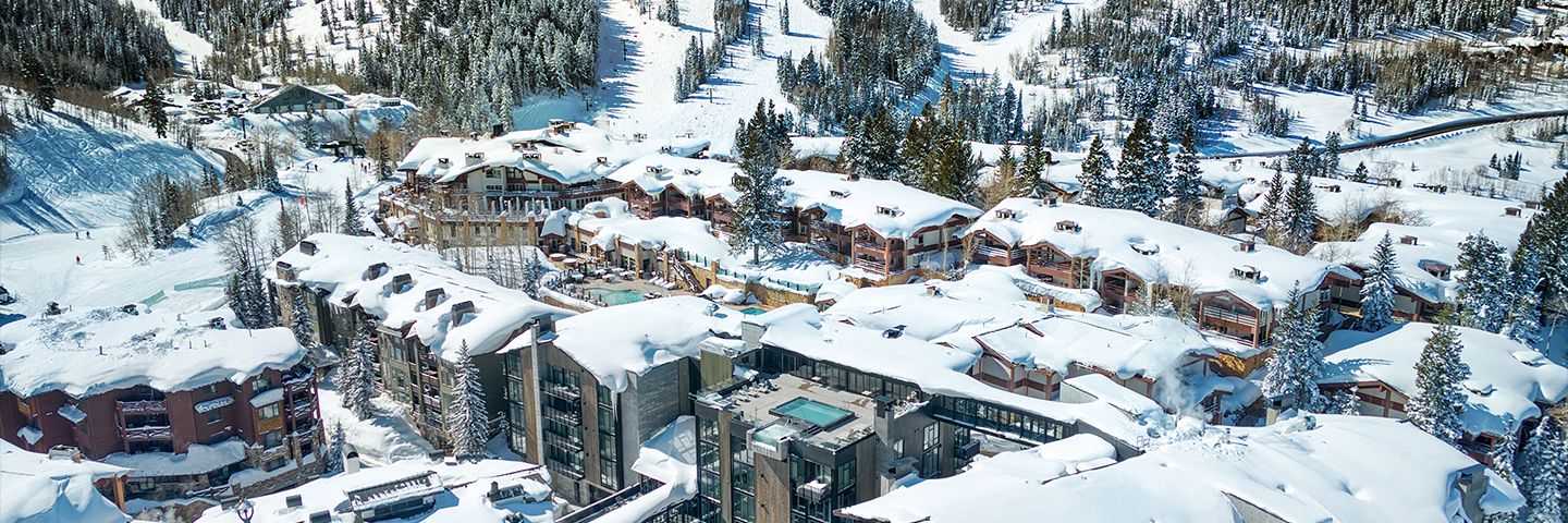 Aerial view of snow-covered luxury lodges at a mountain ski resort surrounded by pine trees.