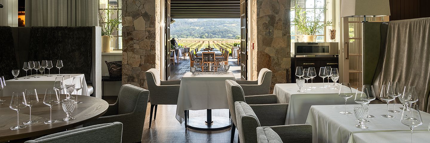 Upscale dining room with stone pillars and set tables opening onto a patio with views of a rolling vineyard landscape.