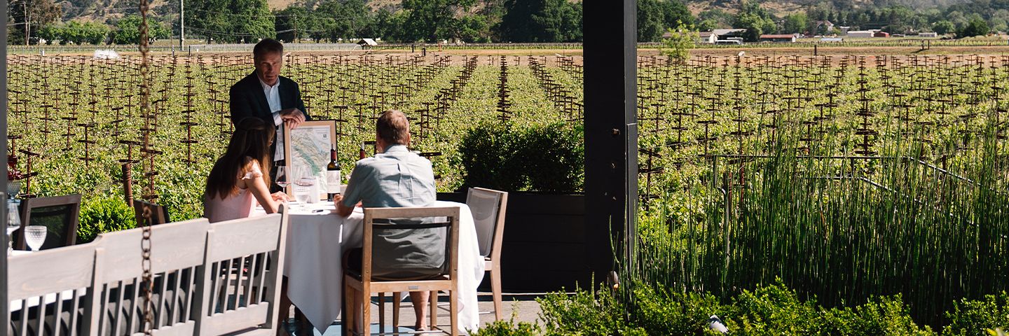 A man shows a map to a couple seated at a table on a terrace overlooking rows of grapevines and distant hills.