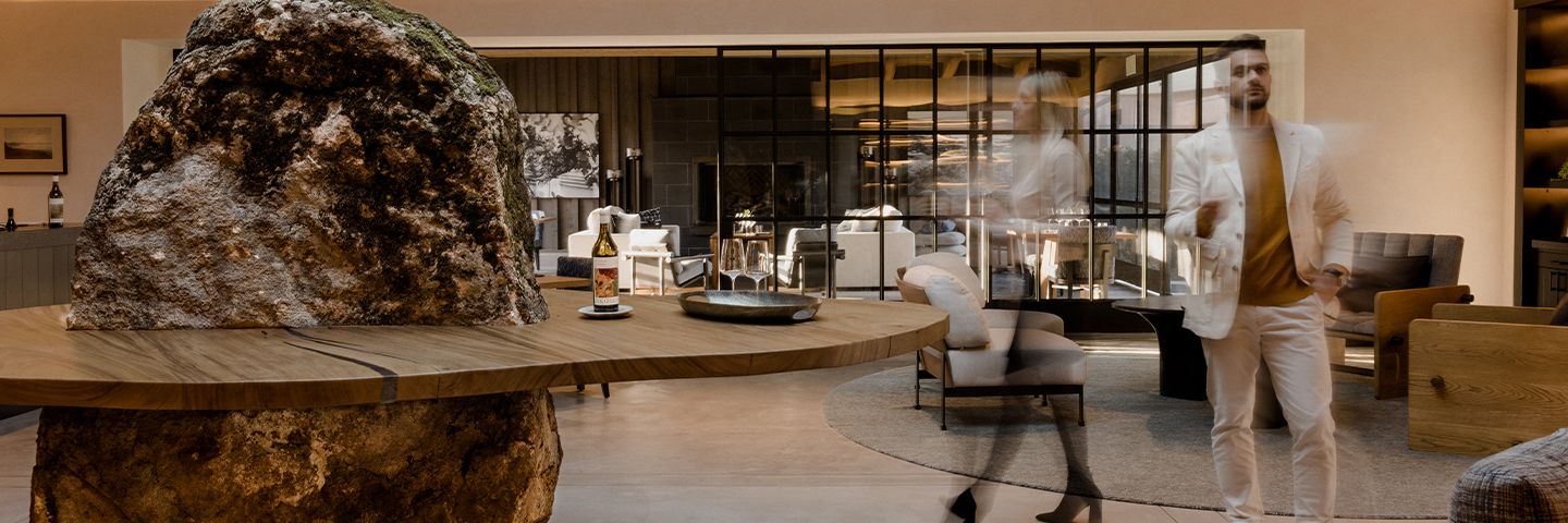 Man in a white suit stands in a luxury lounge featuring a unique rock-base table and motion-blurred figures.