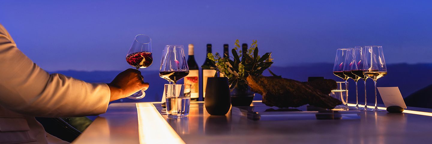 A hand holds a swirling glass of red wine at an illuminated outdoor bar during twilight.