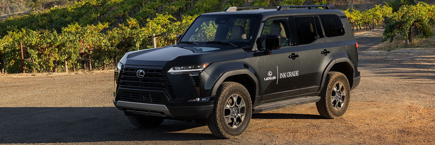 Matte black Lexus SUV with Ink Grade branding parked on a dirt path in front of a hillside vineyard.