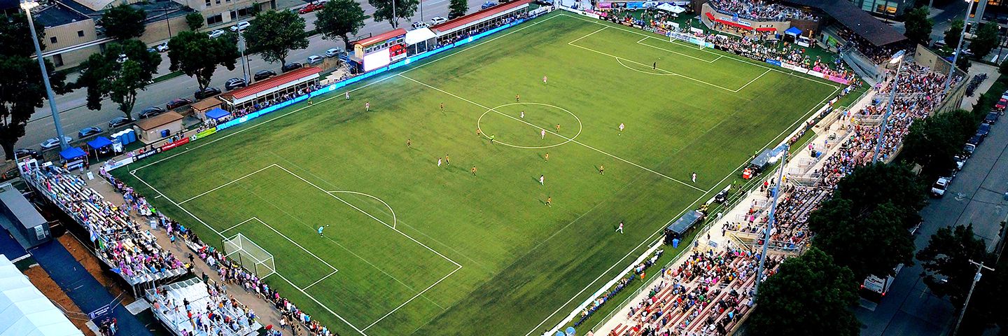 Aerial view of a soccer match in progress at a city stadium with spectators in the bleachers.