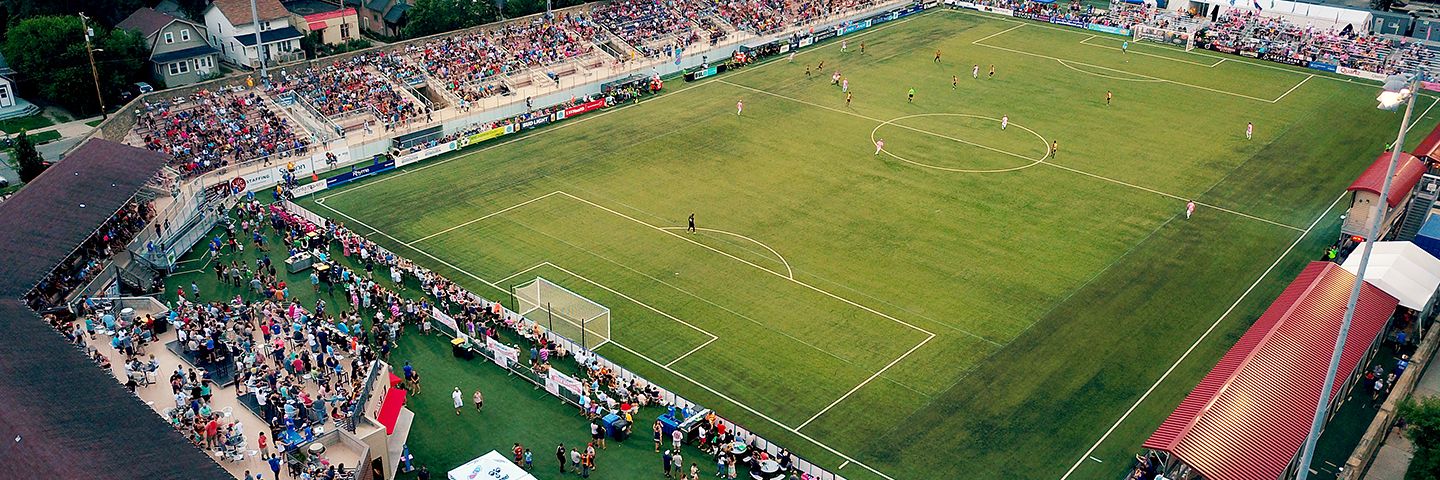 Aerial view of a soccer match in a crowded outdoor stadium at dusk, surrounded by trees and neighborhood houses.