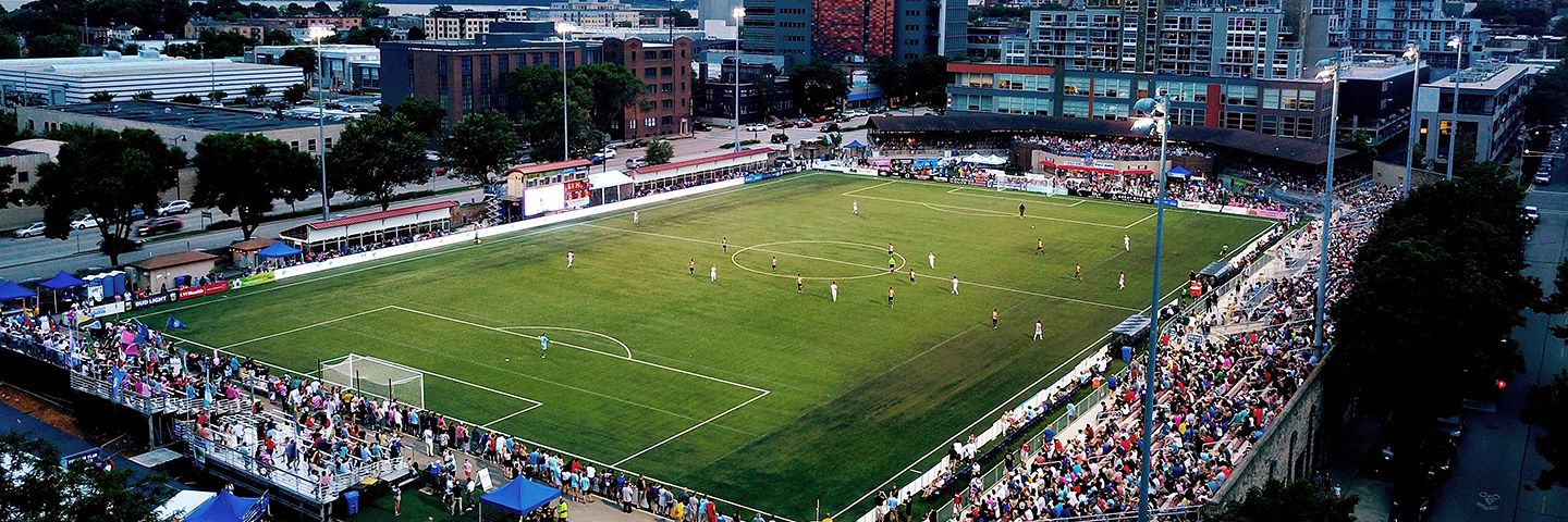 Soccer game at an urban stadium at dusk, with players on the field, spectators in stands, and a city skyline beyond.